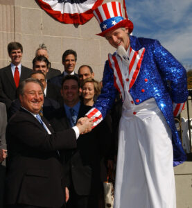 At a corporate Fourth of July event, an event performer on stilts dressed as Uncle Sam shakes hands with a man in a suit. They're stood in front of a group of other smiling people in business attire at the party.