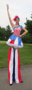For a fourth of July event, a performer on stilts dressed in red, white, and blue with a feathered hat poses with one hand resting as a fist on her hip and another outstretched to the sky.