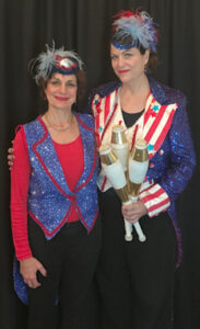 Liz (on right) is dressed in a blue sequined jacket and feathered cap holding gold and white juggling bottles. She is standing next to a woman also dressed in American Flag colors for a Fourth of July event.