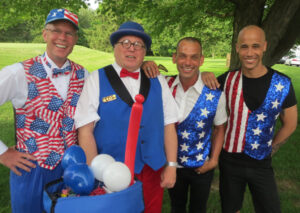Four Independence Day event staff dressed in red, white, and blue, stars & stripes. The second wears a name tag reading "Leo" and he has a box of balloons for balloon sculpting.