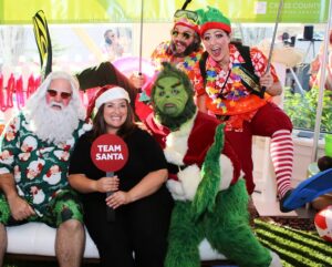 A "green Christmas" party with event performers dressed in Hawaiian tropical shirts. In the front row there is Santa Claus wearing a printed Santa shirt with holiday themed shorts, one dressed in a Santa hat holding a sign that reads "Team Santa", and the Grinch. Behind them are folks dressed as elves in tropical printed shirts and flippers.