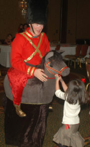 A performer dressed in a costume of a toy soldier riding a magic horse puppet with a child at a Christmas Party