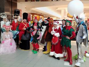 A group of performers dressed in Christmas holiday themed costumes. Costumes include a white reindeer, someone in a flowing pink dress with butterfly wings and pink wig, a nutcracker, a toy soldier on a horse, elves, a snowman, a turkey, Mrs. Claus, Dorothy, and Jack Frost.