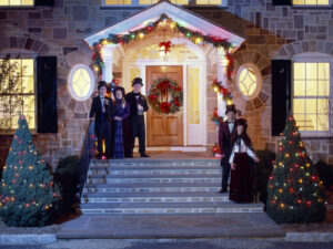 A group of people dressed as Christmas carolers smiling outside a stone house decorated for the holiday party with lights, a wreath, and garland.
