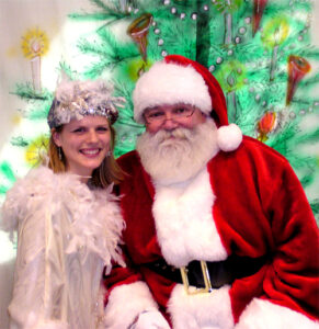 A blonde woman in a white feathered dress and hat with shiny silver accents next to Santa Claus at a holiday event, against a Christmas Tree background.