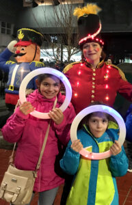 Liz poses with children at an outdoor Christmas party dressed as a toy soldier in lights. The children are posing behind light up juggling rings.