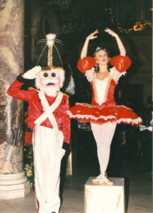 Two Christmas event performers, on the left is one dressed in a Nutcracker costume saluting. On the right is as Clara in a red and white ballerina tutu with arms in fifth position
