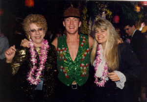 Two women wearing purple leis on either side of a man wearing a sparkly green Christmas vest with candy canes on it at a holiday party.