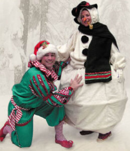 Two Christmas event staff posing, on the left is an elf kneeling next to a snowman standing on the right against a background of snowy trees