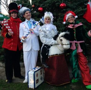 A group of 4 event performers dressed in costumes. From left to right: One in a red and black toy soldier costume holding large candy canes for juggling, one Jack Frost, another on a horse magic puppet, and the last is dressed as an elf, all wearing holiday lights.