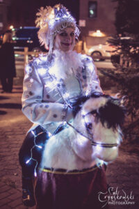 An event performer dressed in white and silver snowflakes, icicles, and lights, on a horse puppet for a Christmas event outside.