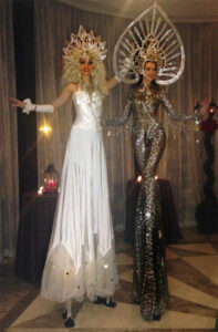 Two performers on stilts dressed in glittery, extravagant costumes for a holiday party.
