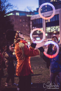 Liz dressed as a toy soldier covered in LED lights juggling light up rings at an outdoor Christmas party.
