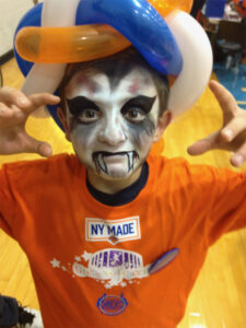 A child in black and white vampire face paint poses with hands outstretched in a creepy pose wearing an orange shirt and blue, white, and orange balloon hat.