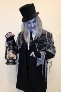 An actor holding a lantern is dressed as the Crypt Keeper in a shoulder-length gray wig, black velvet top hat, and a black suit covered in spiderwebs. Their makeup is done to make their complexion gray and they have a spook grin on their face.
