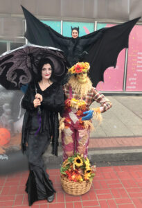 Standing in front of a performer on stilts dressed as a bat with her wings outstretched, are two more event performers. The one on the left is dressed as Morticia Addams holding a black umbrella with an octopus printed on, and on the right is dressed as a scarecrow with a basket of flowers at their feet.