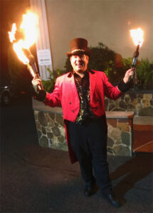 A juggler dressed as a ringmaster smiles while holding up juggling bottles that are on fire.