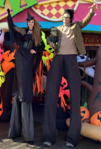 Two event performers on stilts pose in front of blow-up neon ghosts and a spooky tree. The performer on the left is dressed as a witch in all black, while the one on the right is dressed as Frankenstein's Monster.