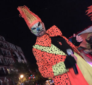 A Halloween event entertainer holding a microphone wears a neon orange & yellow polka-dot clown costume with a "scary clown" mask.