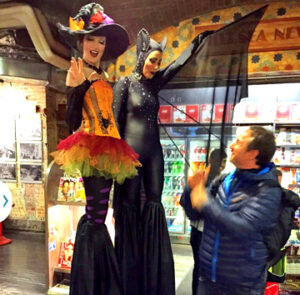 Two event performers on stilts dressed in spooky costumes for a Halloween event pose with an attendee on their right, who is looking up at them. On the left, the performer wears an orange, red, and yellow witch costume with black and purple tights, and the one on the right wears a black bat costume.