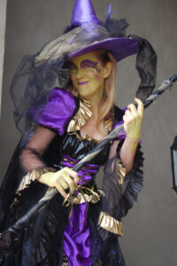 An actress poses against a gray background dressed as a witch in purple and black. Her face is painted green with purple graphic eye makeup while she holds a black and silver broomstick.