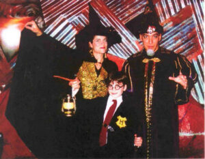 At a Halloween event, Liz is dressed as a witch in mostly black with a gold sequined spiderweb print vest. On her right is a man dressed as a wizard and in front of them is a boy dressed as Harry Potter, holding a wand and lantern.