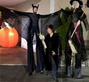 Liz, dressed in black with hints of orange and purple on a feathery hat, poses with gold juggling bottles in between two performers on stilts. On the left the stilt performer is in a black bat costume, on the right the other is dressed as a witch holding a broom.