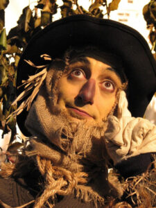 A close up shot of an actor posing with their finger to their temple in a scarecrow costume and makeup.