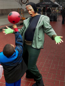 At an outdoor Halloween Party, an actor dressed and made up as Frankenstein's Monster looks surprised as a young boy balances a red ball on his knuckle.